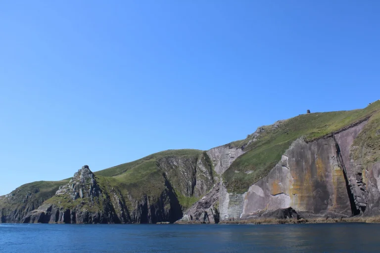 Rocky green cliffs rise from the blue sea under a clear sky, with striations visible on the rock faces and gentle waves at the shore.