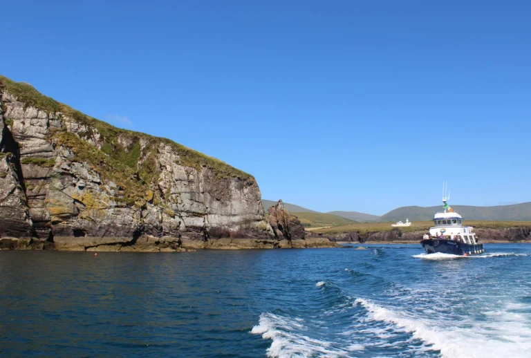 A small boat travels on blue water near rocky cliffs and grassy hills under a clear blue sky.