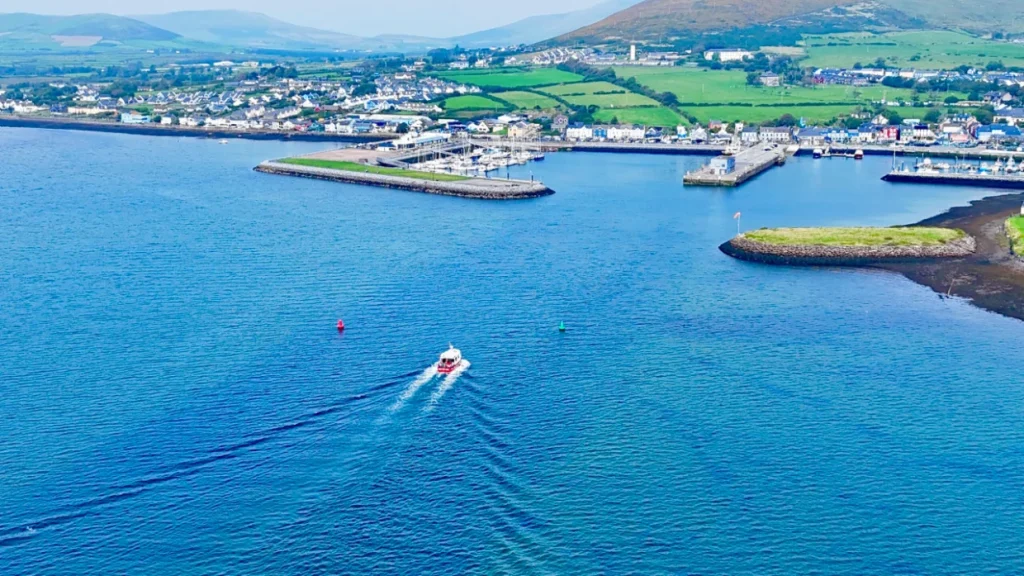A boat travels through the blue water, leaving a wake. In the background, Dingle town sits beside a harbour filled with boats, with green hills beyond.