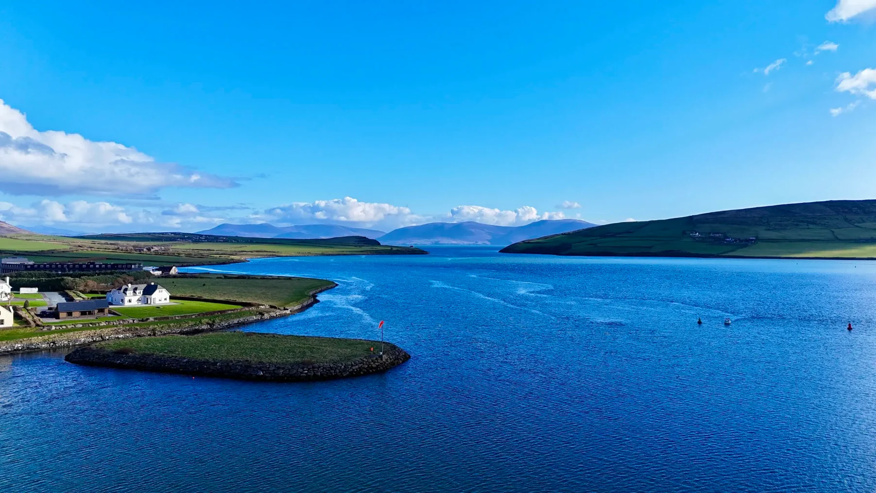 A calm blue bay in County Kerry with a small grassy island, coastal houses on the left, and green hills in the background under a bright blue sky with scattered clouds.