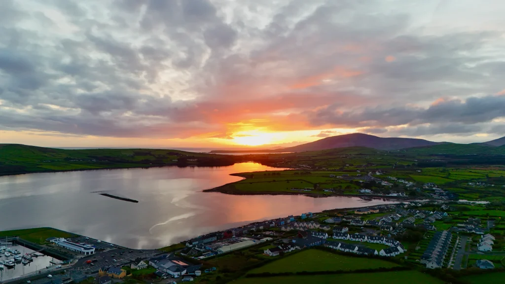 Aerial view of Dingle, a small coastal town in Ireland with houses and green fields by a calm bay at sunset, with hills in the distance under a cloudy sky.