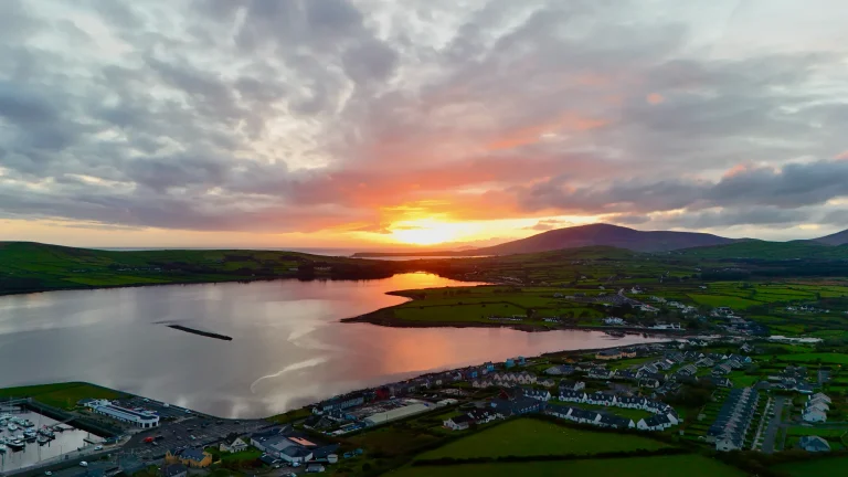 Aerial view of Dingle, a small coastal town in Ireland with houses and green fields by a calm bay at sunset, with hills in the distance under a cloudy sky.