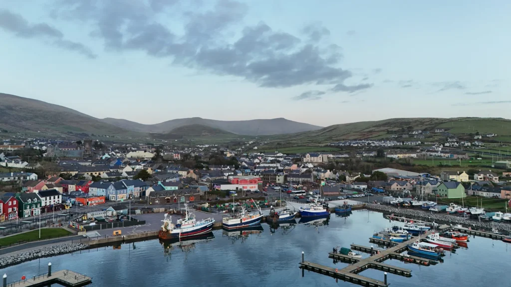 Dingle town with colourful buildings, several boats moored at Dingle Bay, and rolling green hills in the background under a partially cloudy sky. Family day in Dingle