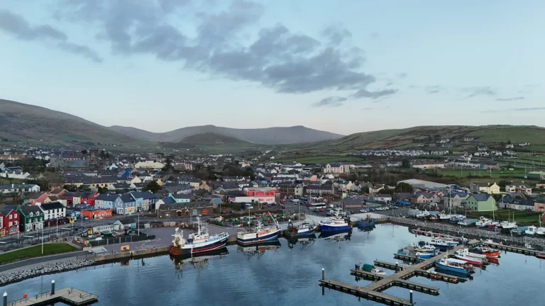 Dingle town with colourful buildings, several boats moored at Dingle Bay, and rolling green hills in the background under a partially cloudy sky. Family day in Dingle